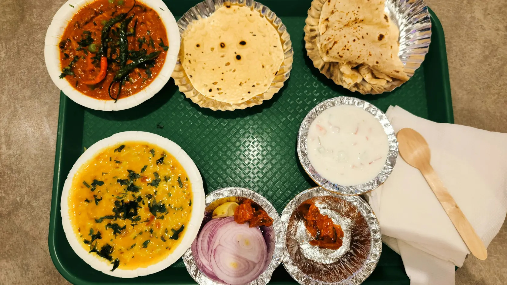 A vegetarian Indian thali on a tray with dal, a curry, rotis, raita, pickle and onion.
